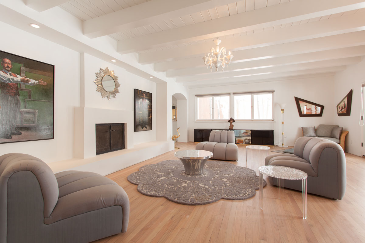 The living room is a spacious sanctuary featuring a gray leather sofa, a center table on a floral cut rug, and a vintage chandelier hanging from the high ceiling. Automated rolling windows and contemporary paintings on the white walls add a fresh, artistic touch to the area.
