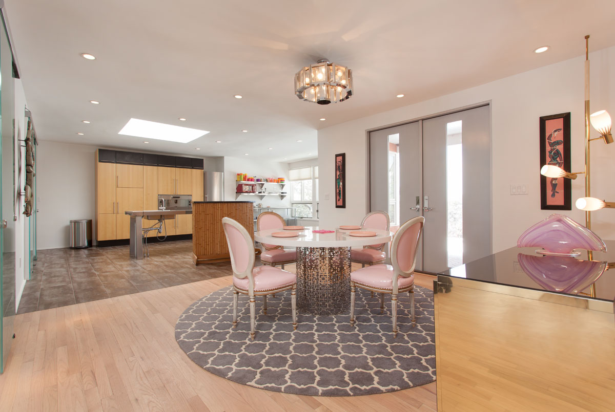 The dining area contrasts with the kitchen's vastness, marked by a chic pink upholstered round dining table and chairs on a geometric gray rug, located near the grand double doors. Modern lighting fixtures in pink, silver, and gold hues, and a minimalist painting, lend an artistic touch to the space.