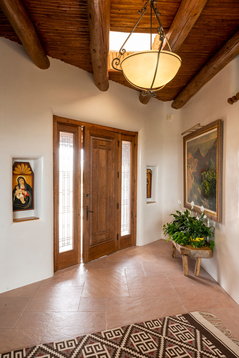 The entrance, marked by rustic wooden doors and flagstone floor tiles, is dramatically enhanced by a skylight and wooden ceiling. A brown and white geometric rug, floral baskets, and a classic painting add to the welcoming atmosphere.