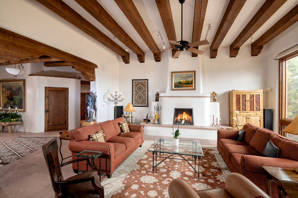 This spacious Southwest-style living room features a brick red sofa, a high white ceiling with wood beams, a central fireplace, and a geometric rug, all illuminated by light streaming through large wooden-framed windows. Flagstone flooring, candle holders, wooden doors, cabinets, and tastefully selected wall paintings add to the room's rustic charm.