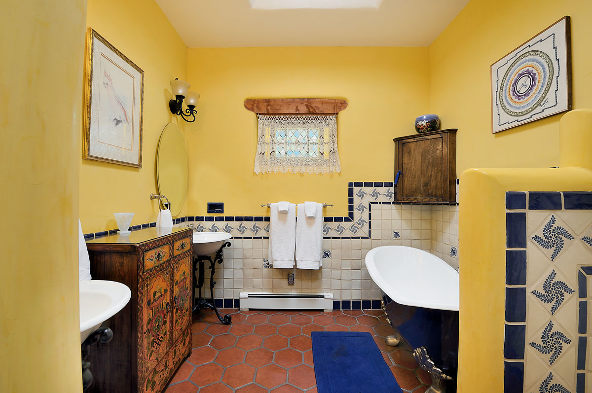 This bathroom boasts a vibrant theme of yellow walls mixed with blue and white tiles, and a matching bathtub. The space features wooden cabinets, a crochet curtain, a blue rug, and two wall paintings, adding charm to the room.