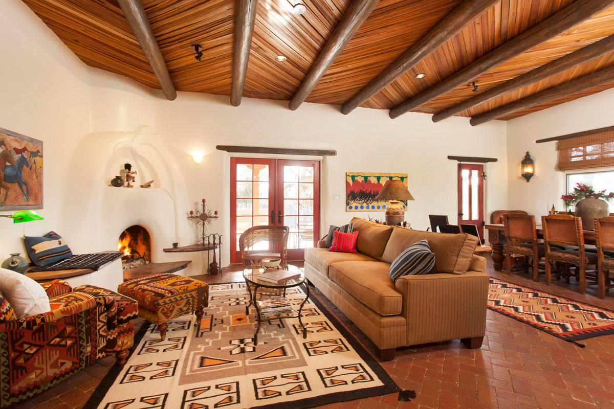 The living and dining rooms, with a rustic Spanish theme, feature wooden furniture, a southwestern sofa, and carpet. Sunlight from French doors highlights the Granada tiles and kiva fireplace against the white walls.
