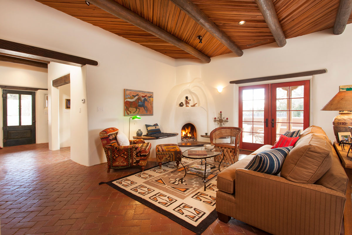 This rustic Spanish-themed living room renovation boasts an ethnic southwest sofa and matching carpet, a traditional kiva fireplace, and French doors that bathe the room in natural light. The unique Granada floor tiles and wood ceiling tiles add to the room's authenticity, creating a harmonious blend of rustic elegance and Spanish charm.