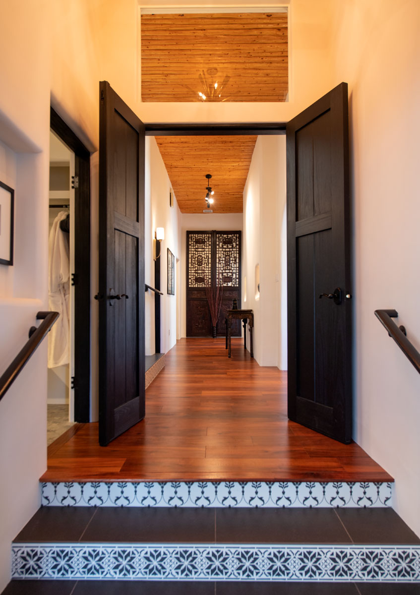 In a fully remodeled home blending Asian design with traditional New Mexico style, a vintage hallway with polished hardwood floors leads to a dark brown wooden door.