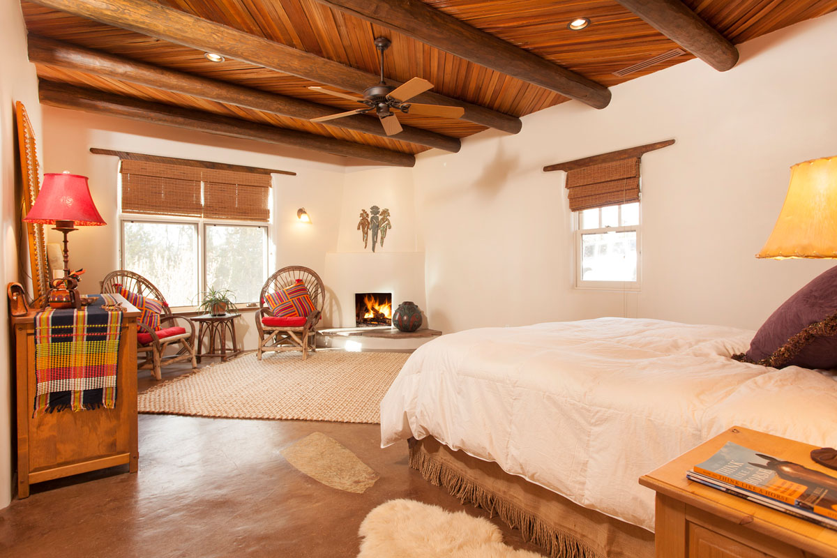 Embracing a rustic Spanish theme, the master suite renovation combines bamboo blinds and wood ceilings for a natural feel, set against warm beige walls that highlight the cozy fireplace. The suite is completed by rattan furniture, contributing to the room's comfortable elegance and rustic charm.