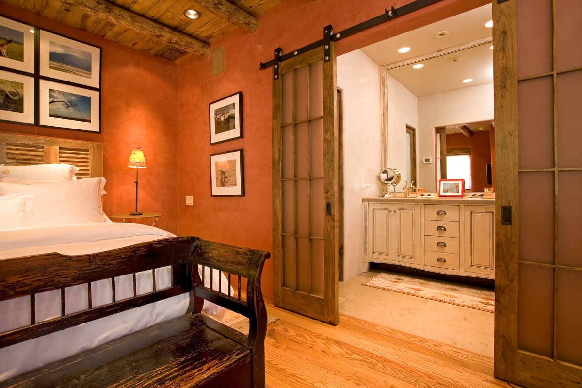 A cozy bedroom features a wooden bed frame, antique bench, and vintage cabinet. Overhead wooden beams add character, and a rustic French barn door leads to the walk-in closet. The room is lit with soft, neutral lighting.
