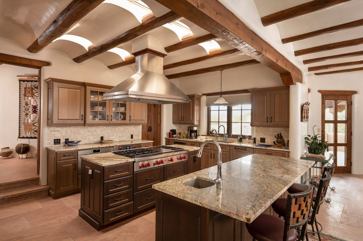 The kitchen radiates elegance with its beige walls, wood beams, and cabinets, highlighted by an island with a granite countertop, red and brown high chairs, and stove burners. The white concave ceiling adorned with dark wooden beams enhances the room's elegant atmosphere.