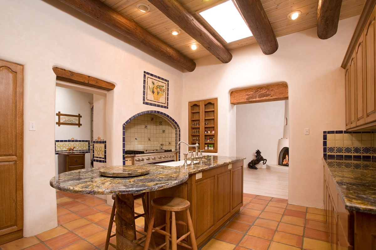 This kitchen blends rustic charm with modern luxury, featuring brown floor tiles, white walls, wood beams, and a skylight. A granite-topped island and table, an oval-tiled stove, and an open door revealing a glimpse of a kiva fireplace in the living room complete the inviting ensemble.