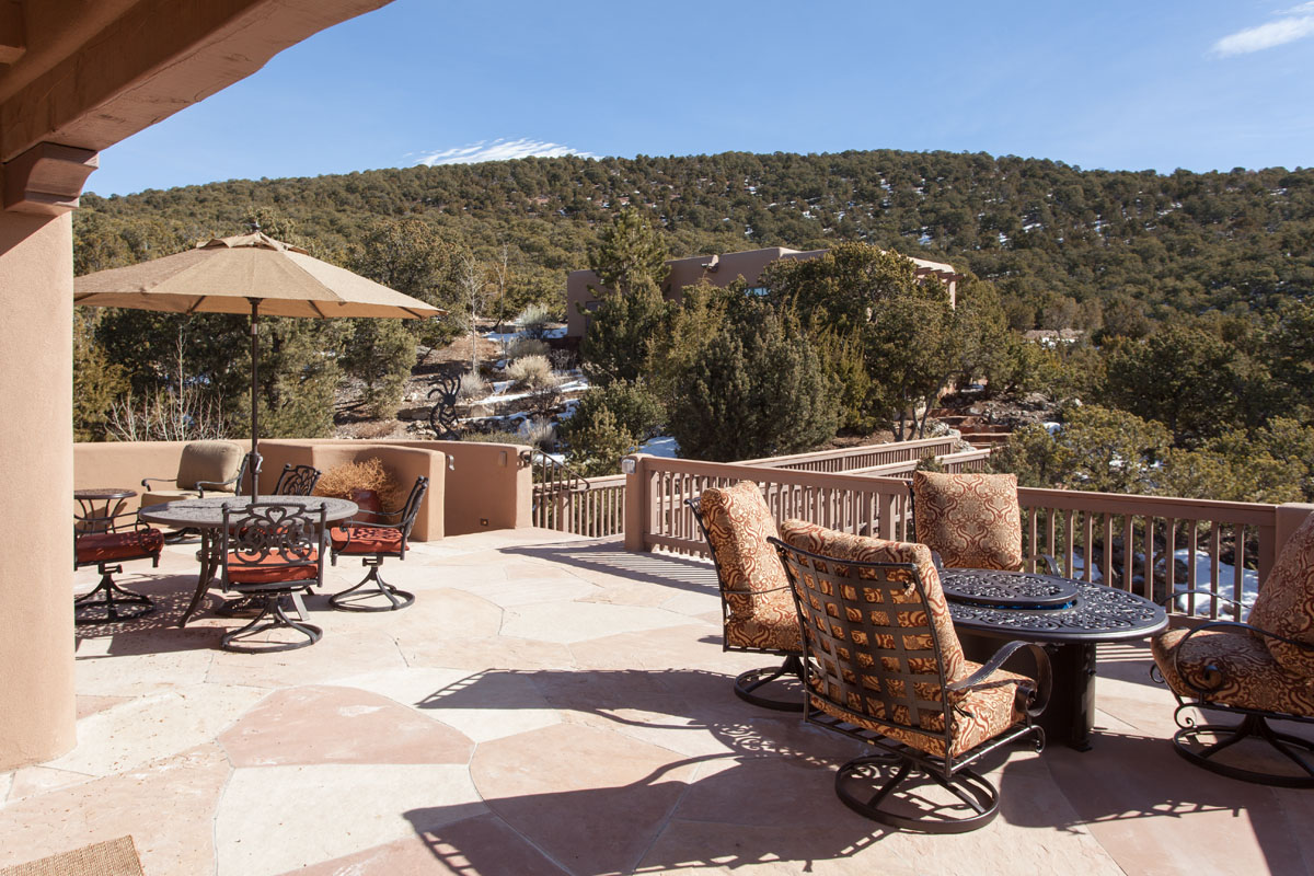 The outdoor patio, with its rustic adobe floor and balusters, is furnished with round tables and chairs under an umbrella. This inviting space offers breathtaking views of the surrounding green trees and distant mountains.