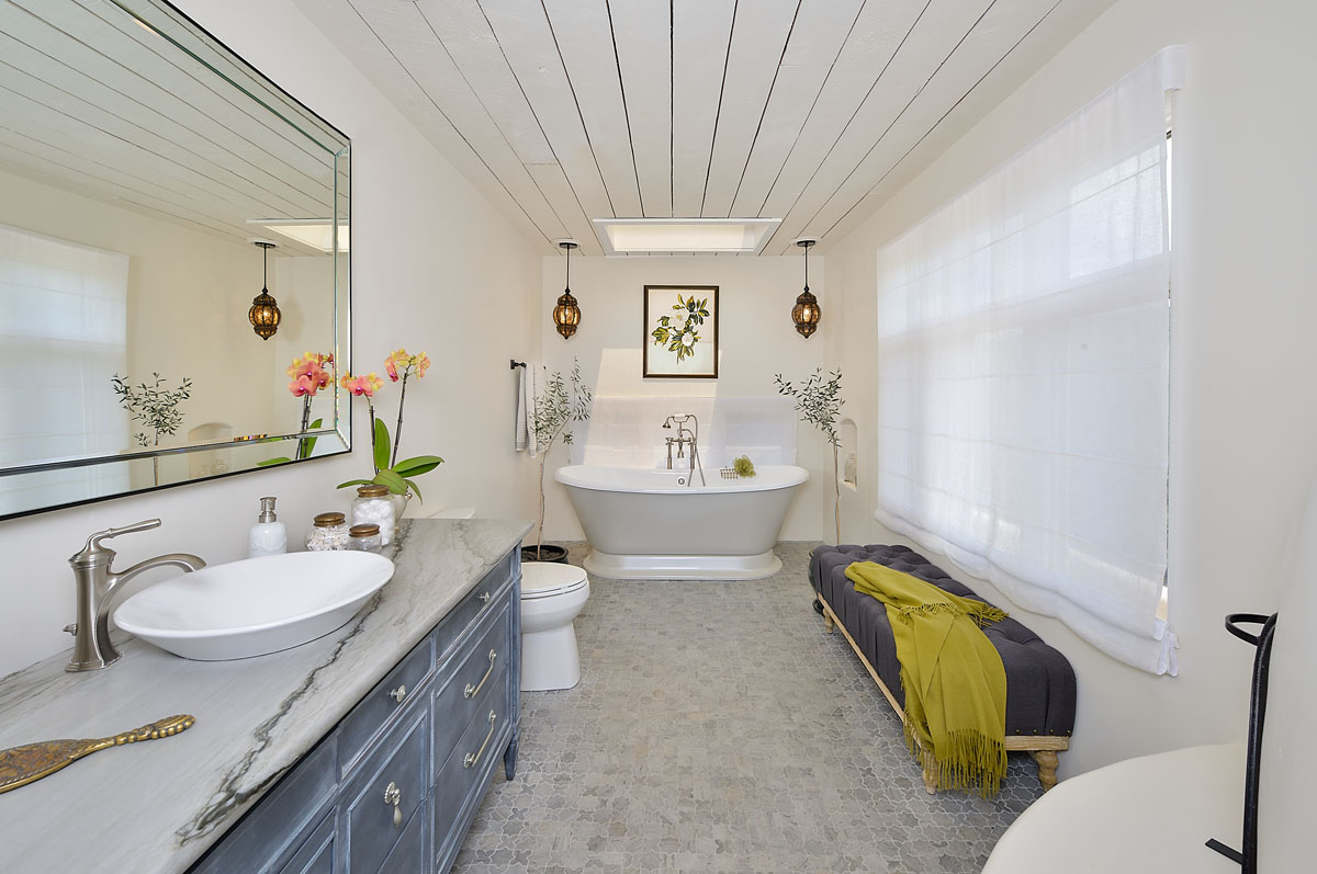 The bathroom showcases a signature marble countertop, a white vintage tub, ambient Moroccan lamps, and a plush velvet ottoman.