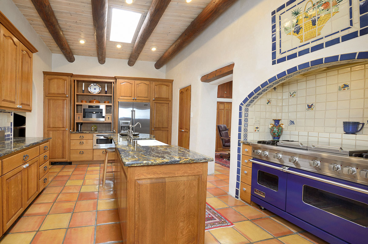 The kitchen showcases a blue-toned built-in stove and oven, set against conclave tiles and topped with a yellow garden pot tile mosaic. A mix of white and wood themes prevails in the cabinetry and central island, which is crowned with a sleek granite countertop.