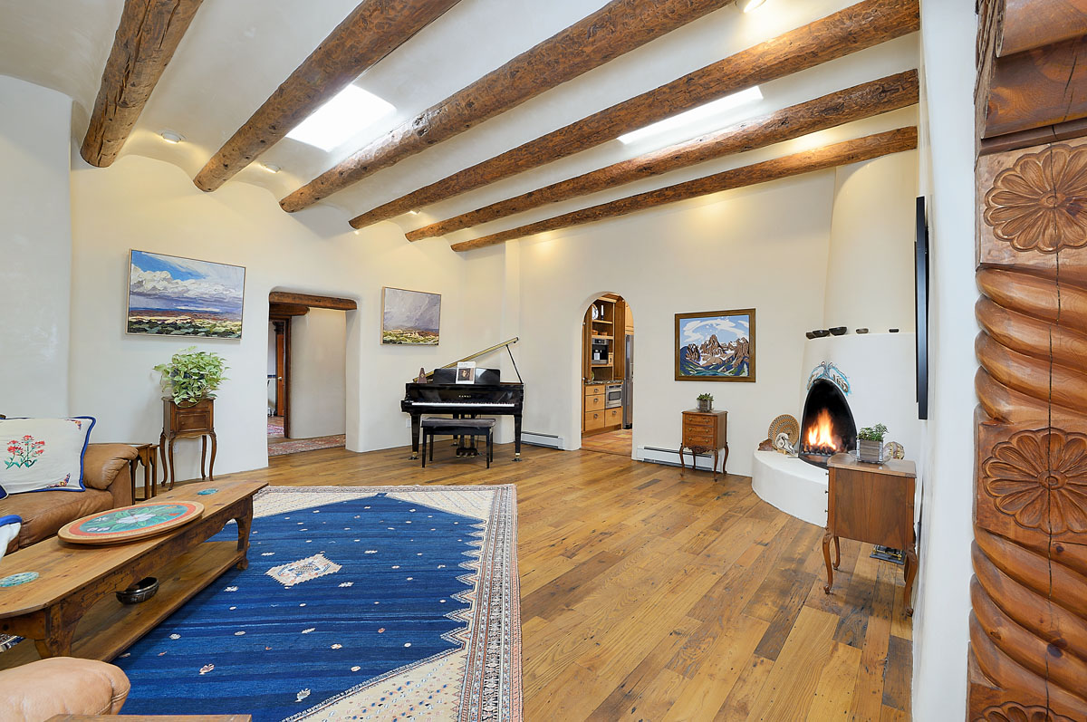 A harmonious blend of old and new, this living room features warm wood floors, white walls with wooden decor, a mocha leather sofa, and a vintage table atop a spacious blue and gray rug. The room's heart is a kiva fireplace, set against a backdrop of a grand black piano, contrasting wood beams, white ceiling, and modern paintings.