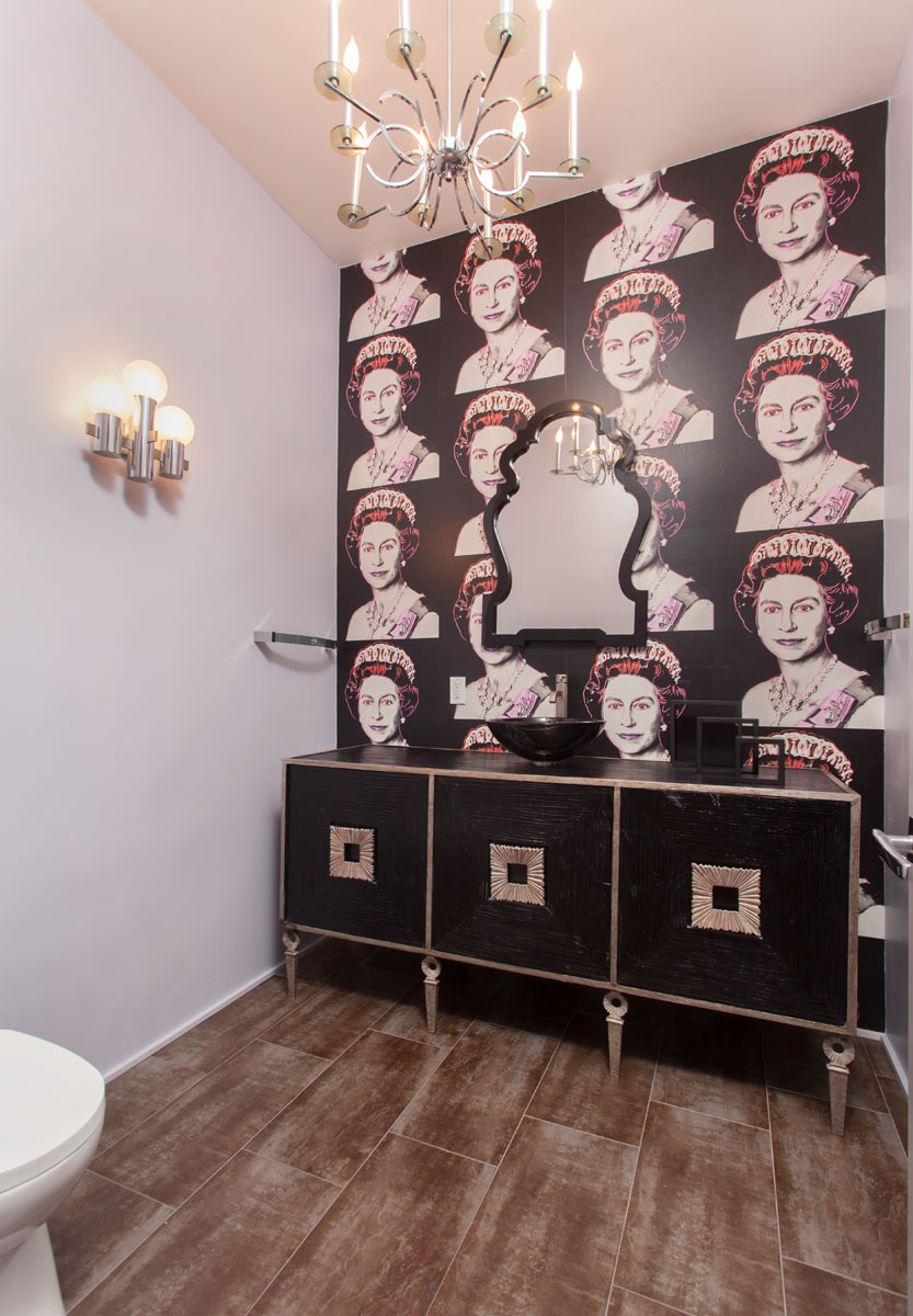 The guest bathroom stands out with its unique black wallpaper featuring a Queen Elizabeth print. The room is further enhanced by a sleek black faucet, three cabinets with white geometric handles, and a chic chandelier that adds a touch of elegance.