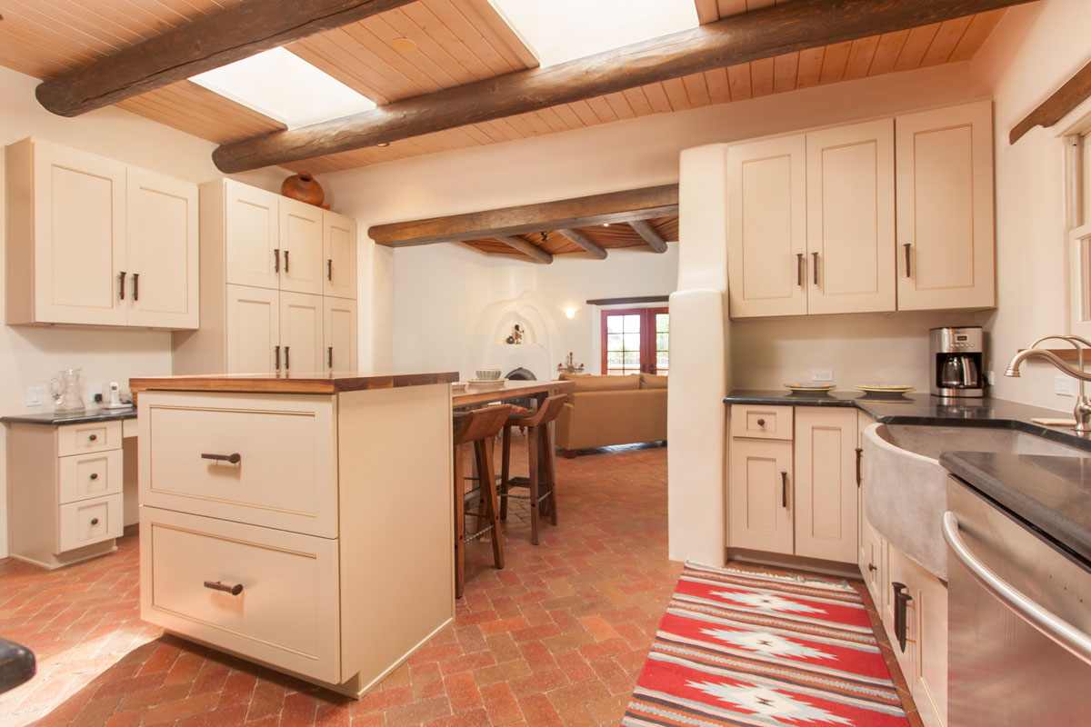 In the kitchen, white oak cabinets and high stool chairs contrast with the white walls, while a gray mid-age oval faucet adds a vintage touch. The skylight, framed by a wood beam, enhances the rustic Spanish charm with natural light.
