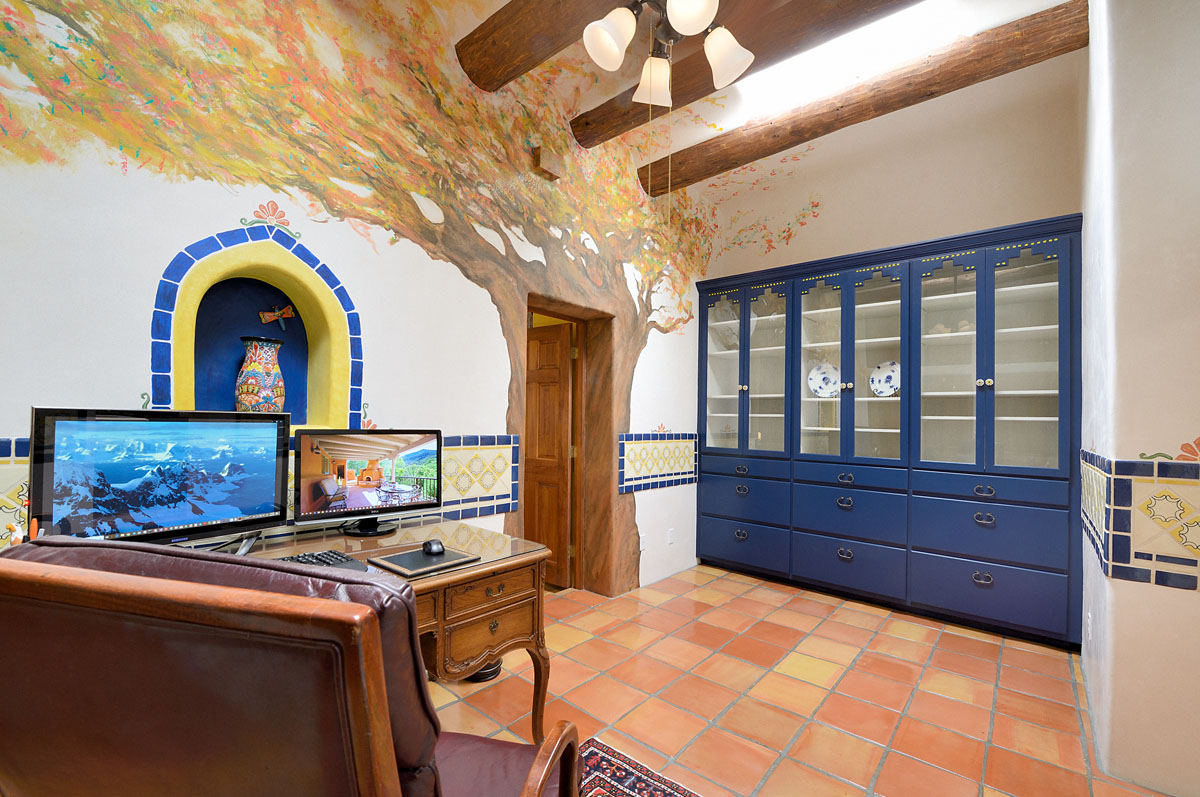 The room features blue cupboards and cabinets against the warm wood-toned floor tiles and includes a corner with a vintage desk setup. The white walls are artistically adorned with a mural of an autumn tree and an engraved blue and yellow wall decoration.
