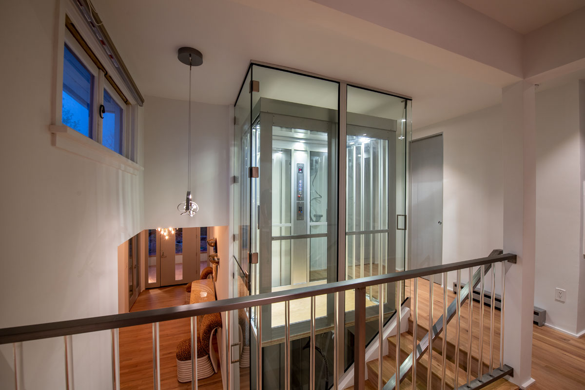 The end of the wooden hallway reveals the house's standout feature: a modern platform lift, made of glass and metal, positioned beside the staircase. An oversized pendant light hanging from the ceiling enhances this unique attraction.
