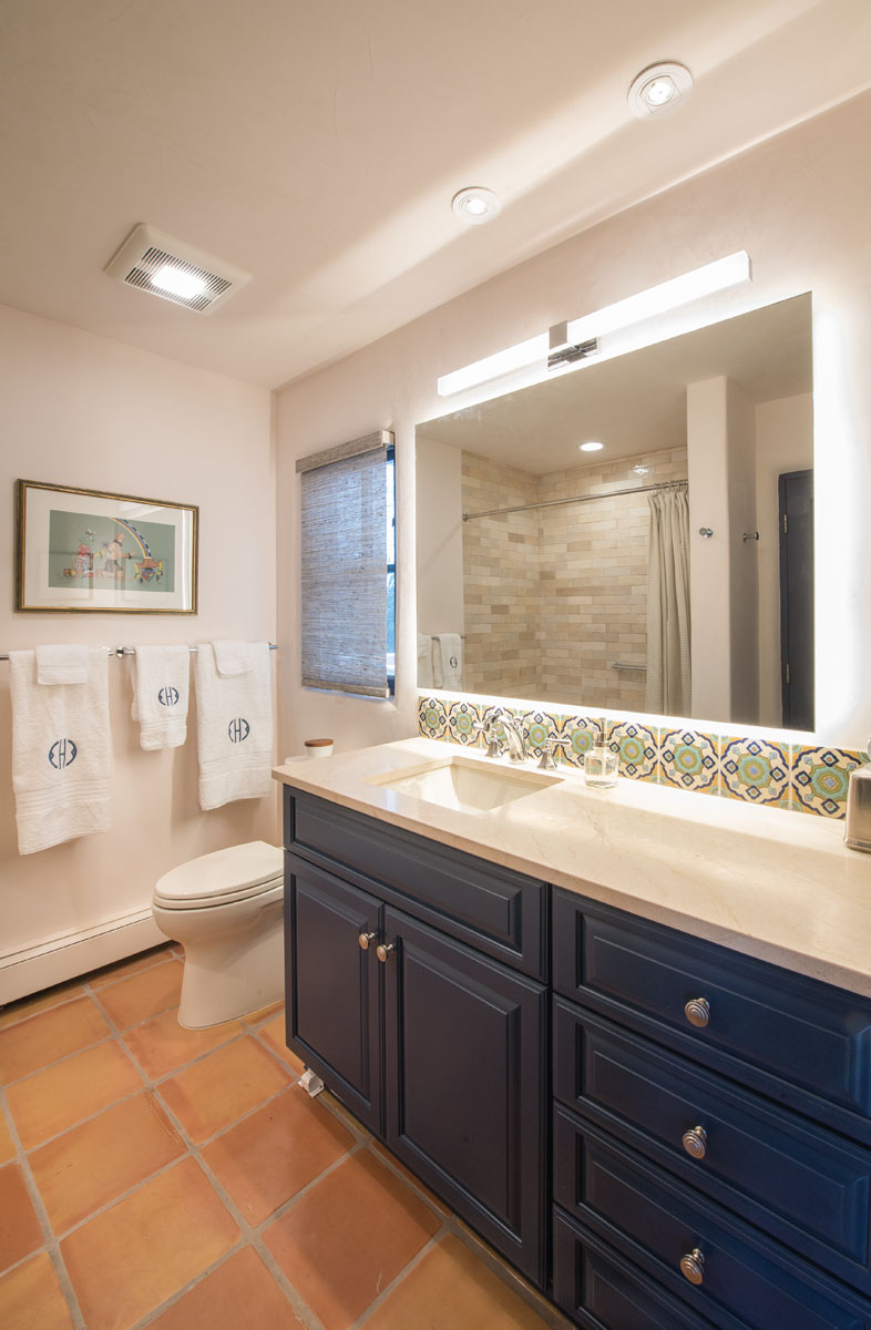 The comfort room combines elegance and simplicity, featuring a navy blue cabinet, clear glass shower door, and plush Weezie towels hung near the toilet bowl, all contrasted against pristine white walls. The white ceiling with a light-fitted fan and two LED lamps illuminate the space, while the warm Saltillo tiles ground the room's aesthetic.