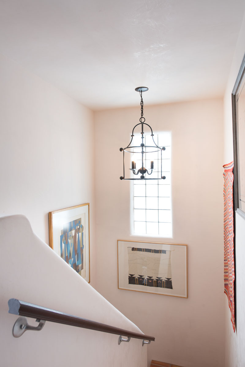 The stairway, adorned with a warm wood handrail and lit by a lantern pendant light, is set against a backdrop of peachy pink walls. A clear glass window invites natural light, further enhancing the colors of the two framed paintings that add a touch of artistry to the ascent.