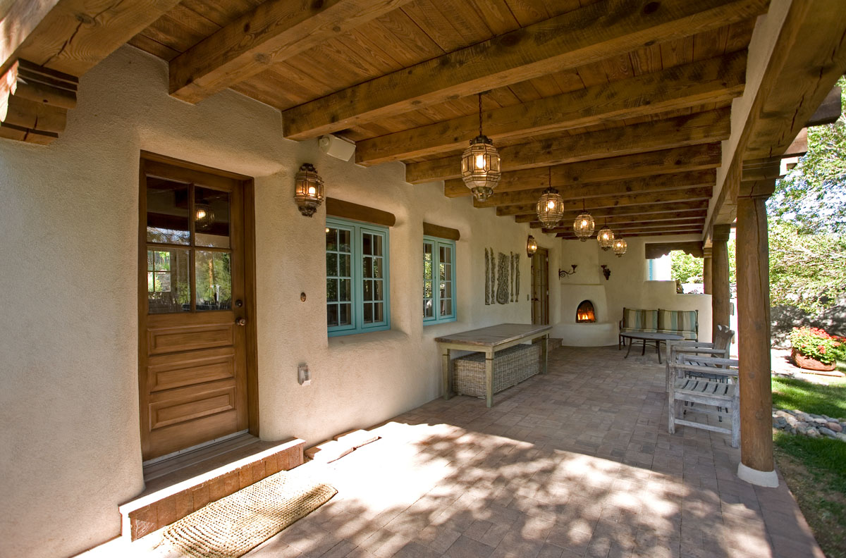 Blending Sante Fe and Spanish themes, the outdoor patio renovation features a kiva fireplace centerpiece and slated gray armchairs for comfort and style. The rustic chandeliers, wood doors, and French blue windows create an enchanting space that radiates warmth and Spanish charm.