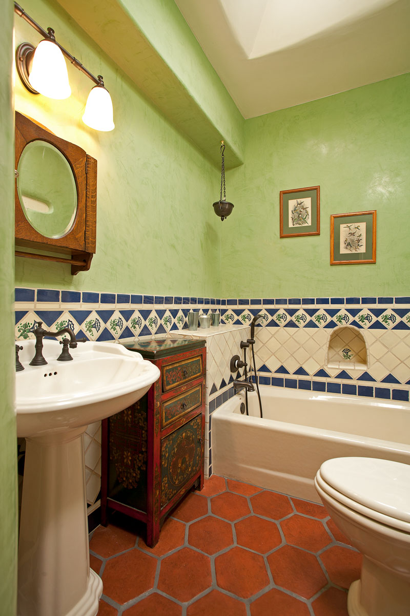 The bathroom combines vintage charm and modern style with its mint green walls, terra cotta floor tiles, and a red wood cabinet next to the lavatory. Hand-painted blue wall tiles and minimalist paintings add artistic touches to the space.