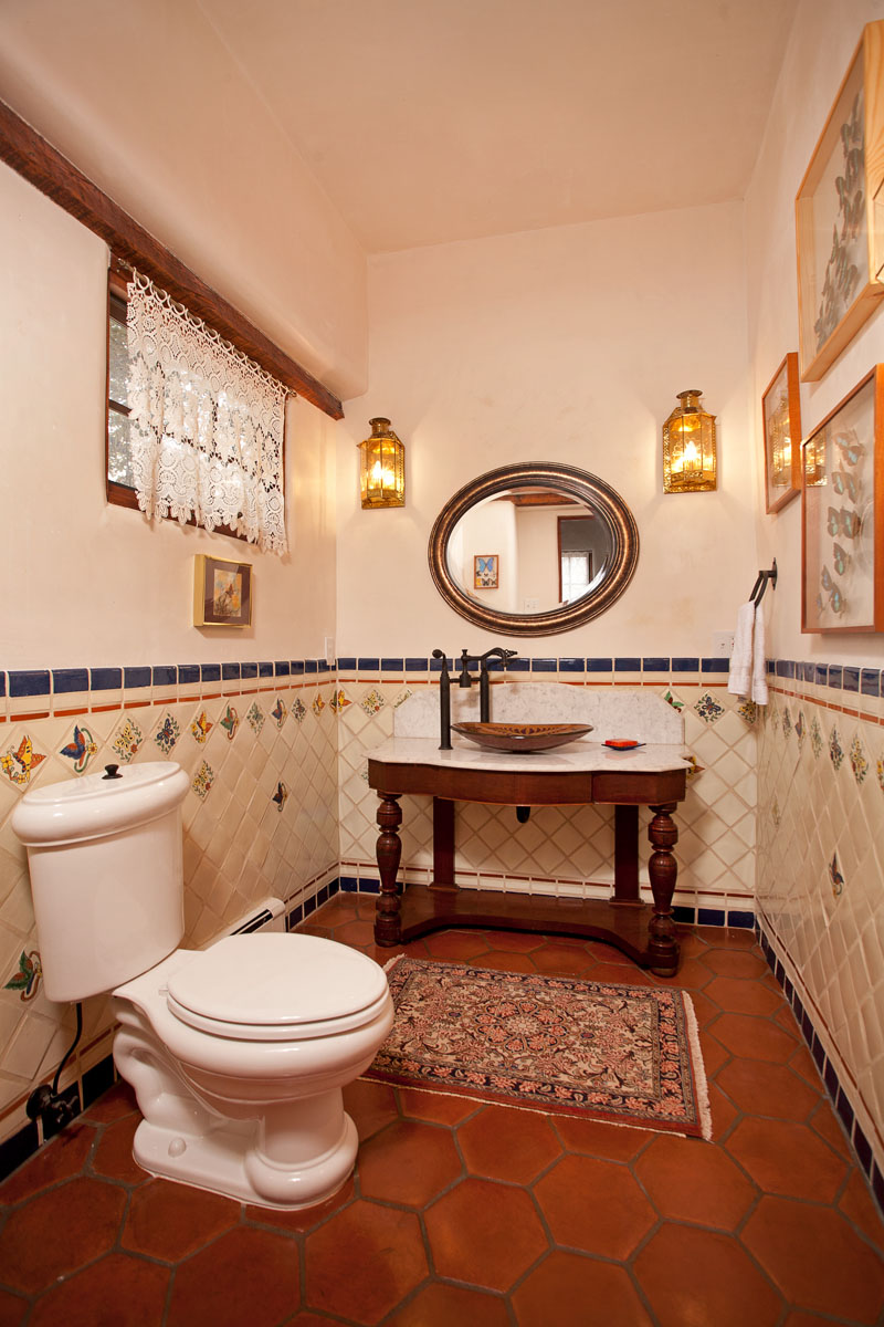 The bathroom exudes classic elegance with its red hexagonal floor tiles, hand-painted blue and cream wall tiles, and a wooden porcelain lavatory. Additional features include a white toilet bowl, red rug, butterfly decoration, lamp, and a white crochet curtain.