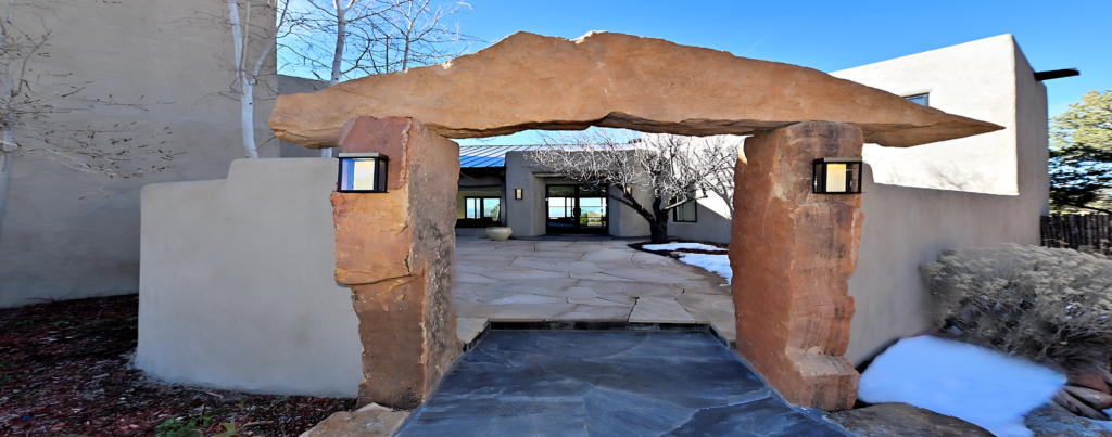 A panoramic view of a southwestern-style entryway featuring a wide stone arch between two plastered walls, with mounted lights and a snowy ground leading to a courtyard.