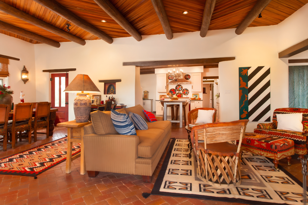 A vibrant living space featuring exposed wooden ceiling beams, colorful rugs, and Southwestern-inspired decor. The room includes a cozy seating area with a mix of woven and upholstered furniture, creating a warm, inviting atmosphere with eclectic design elements.