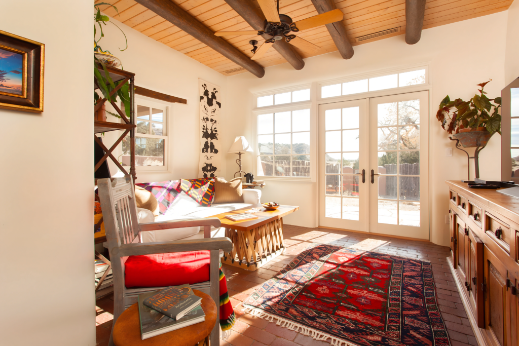 A cozy, sunlit room featuring a large red rug, wooden ceiling beams, and glass doors that let in natural light. The space is decorated with comfortable seating, vibrant colors, and cultural art, creating an inviting atmosphere for relaxation and entertainment.