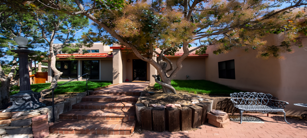The image showcases a well-landscaped exterior view of a house, featuring stone steps leading to the entrance, lush greenery, and a decorative bench. The architecture reflects a harmonious blend with the surrounding nature, highlighted by a tree in the foreground.