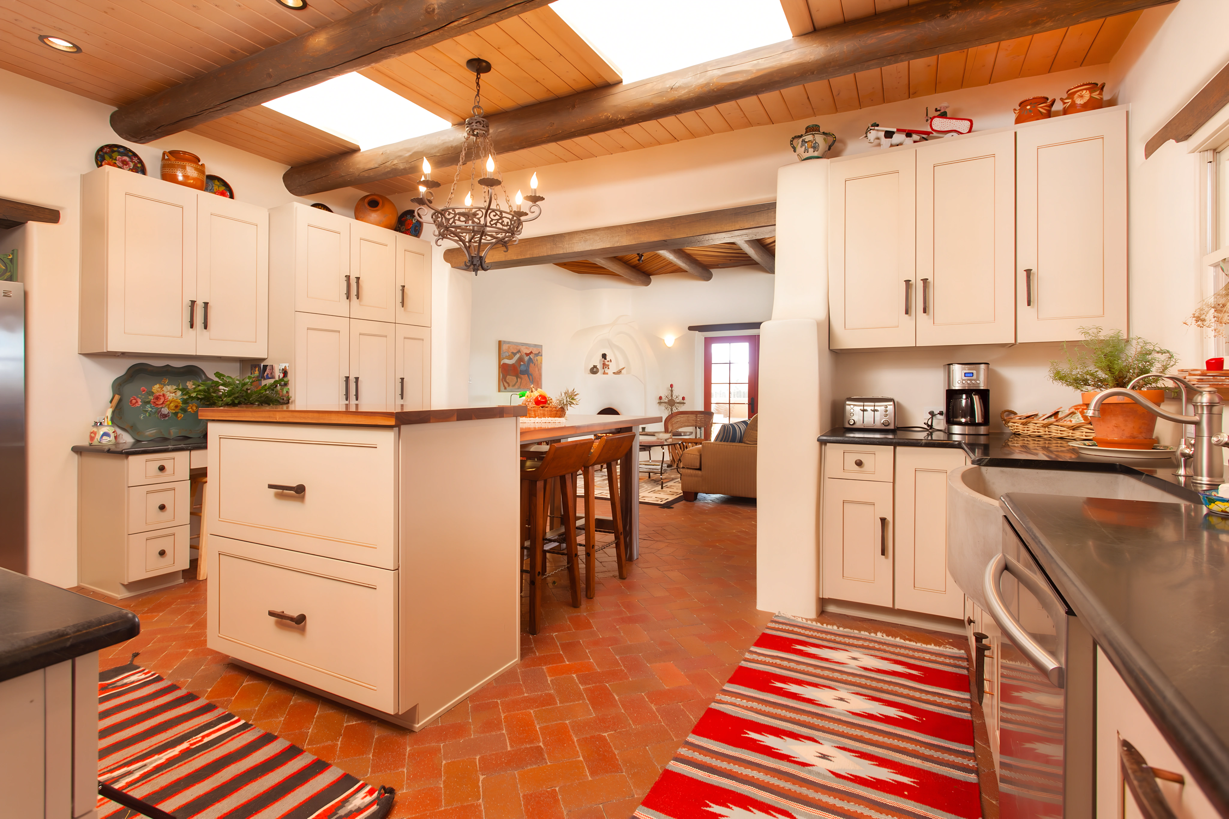 A charming Southwestern-style kitchen with terracotta tile floors, white cabinetry, and exposed wooden beams. The space is adorned with colorful rugs, a central island, and a chandelier, creating a warm, inviting atmosphere for cooking and dining.