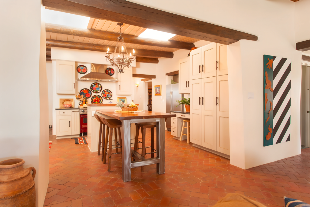 A beautifully renovated kitchen featuring terracotta tile floors, white cabinets, and a central wooden island. The space is adorned with colorful plates, a chandelier, and exposed wooden beams, creating a warm, inviting, Southwestern-inspired atmosphere.