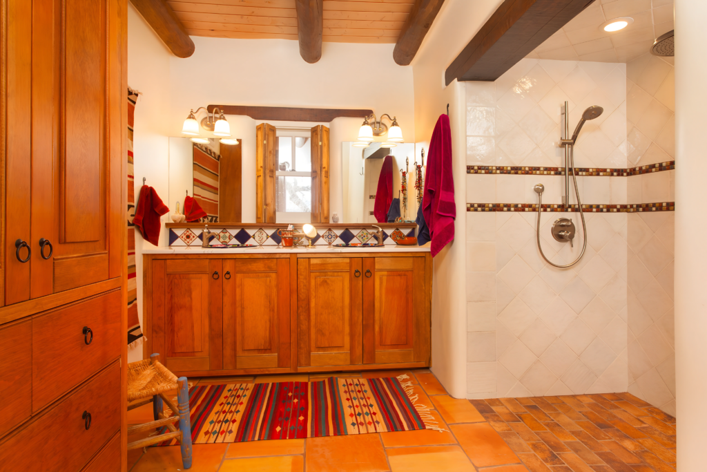 A warm, Southwestern-inspired bathroom featuring terracotta tile floors, wooden cabinets, and a walk-in shower with decorative tile accents. Exposed wooden beams add rustic charm, while colorful rugs provide a cozy, vibrant touch to the inviting space.