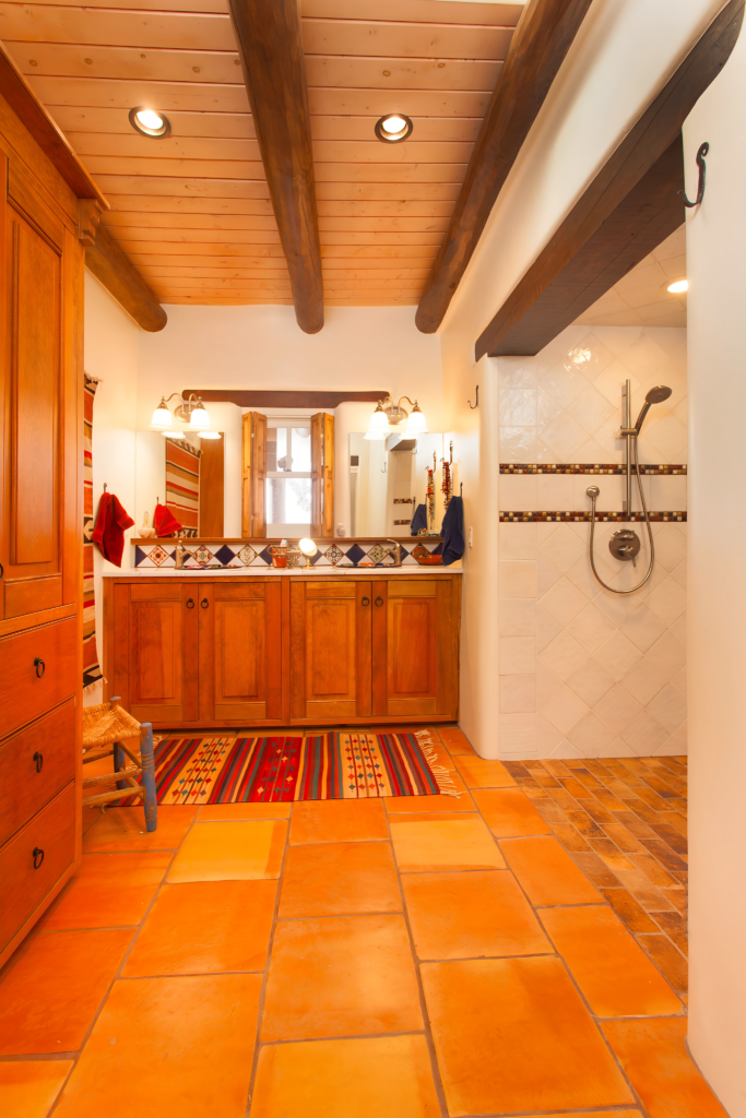 A beautifully renovated bathroom featuring warm terracotta tiles, wooden cabinetry, and a spacious walk-in shower with elegant tile work. The ceiling showcases exposed wooden beams, while colorful rugs add a vibrant, cozy touch to the space.