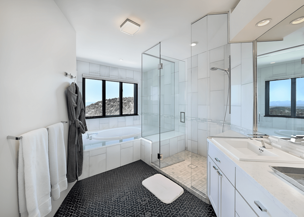 A contemporary bathroom featuring a glass-enclosed shower and a freestanding soaking tub beside large windows that offer panoramic views. The space has sleek white cabinetry, modern fixtures, and dark textured flooring, creating a stylish and relaxing atmosphere.