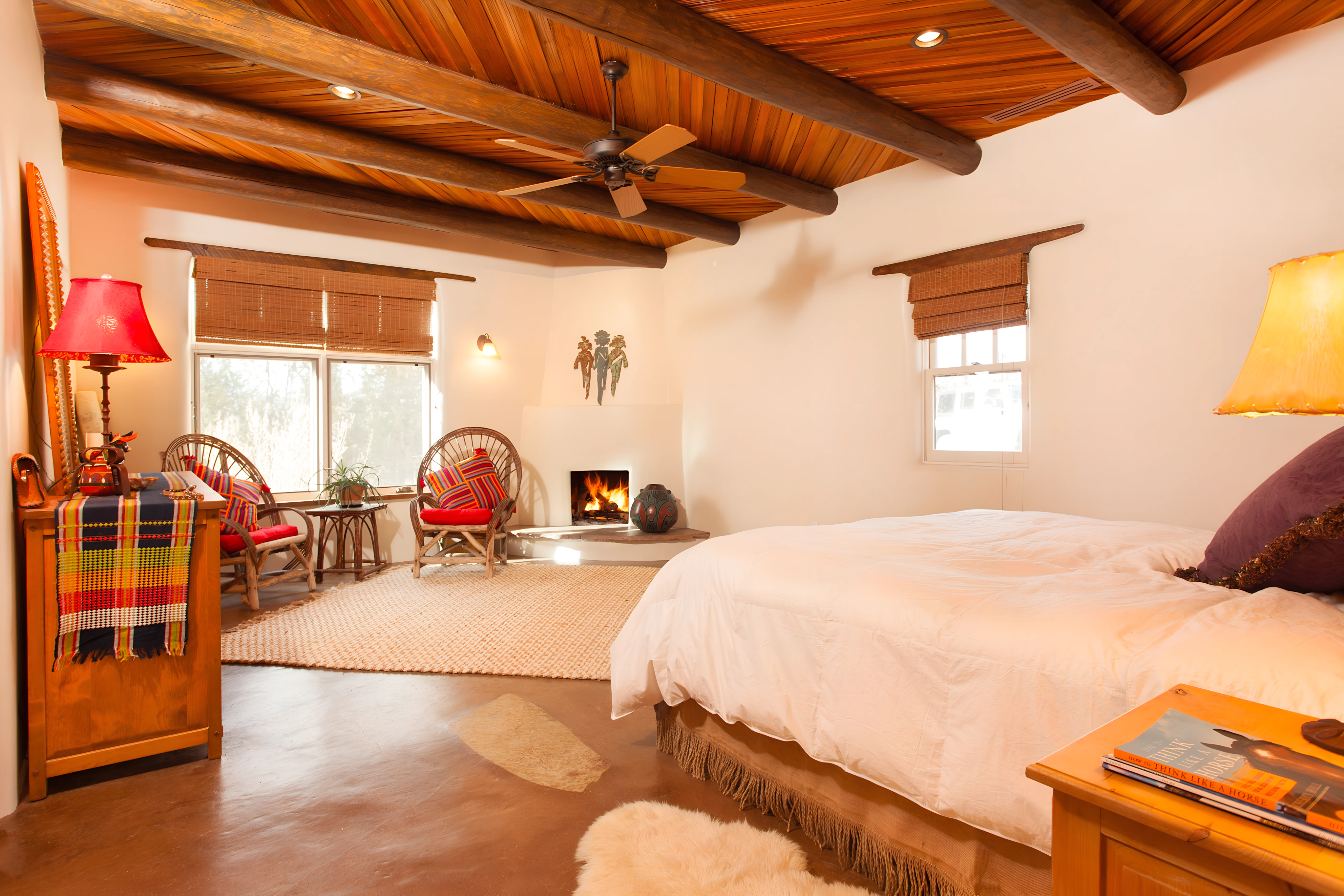 The image shows a beautifully decorated bedroom with a warm and inviting atmosphere. The room features a cozy bed with soft bedding, a fireplace with an artistic clay pot beside it, and two comfortable chairs arranged around a window with views of the outdoors. The ceiling is adorned with exposed beams, and the room is illuminated with ambient lighting from a bedside lamp and wall sconces. The room's earthy tones, warm lighting, and rustic furniture create a peaceful retreat.