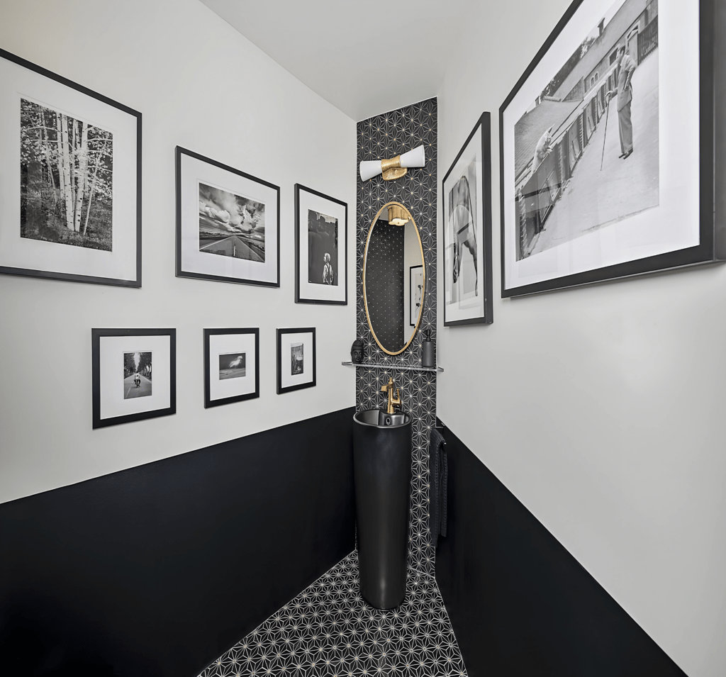 A chic powder room featuring a unique design with black and white elements. The space showcases a modern pedestal sink and a large oval mirror, surrounded by framed black-and-white photographs. The patterned floor adds a stylish touch to the contemporary decor.