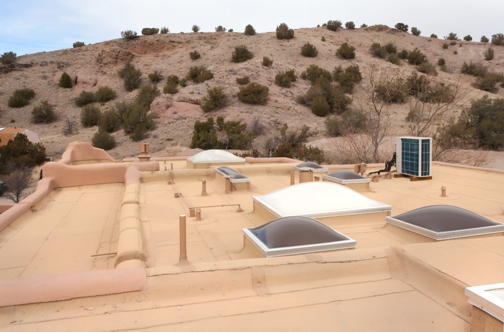 A flat, tan-colored rooftop with multiple skylights and an air conditioning unit sits against a backdrop of desert hills and scattered vegetation. The clean and finished surface contrasts with the natural landscape, suggesting completed renovation work.