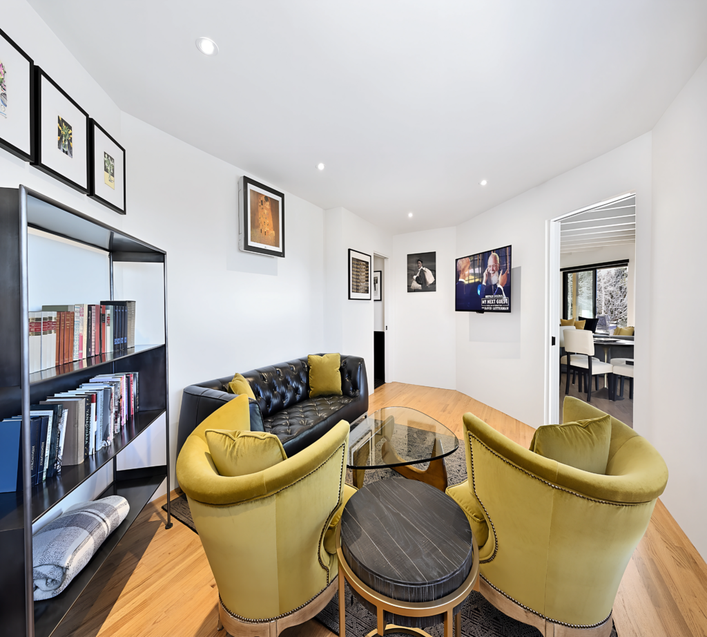 A modern TV room featuring a sleek design with a black bookshelf filled with books. Two vibrant yellow chairs surround a glass coffee table, creating a cozy seating area. Framed artwork decorates the walls, and large windows provide natural light.