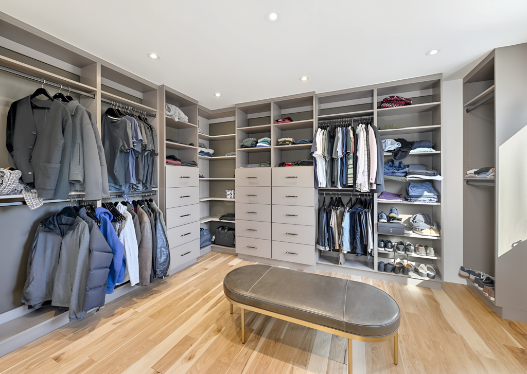 A stylish walk-in closet featuring organized shelves filled with folded clothes and hanging garments. A central bench adds a practical touch, while warm wooden flooring and bright lighting create an inviting atmosphere. Drawers line one wall for additional storage.