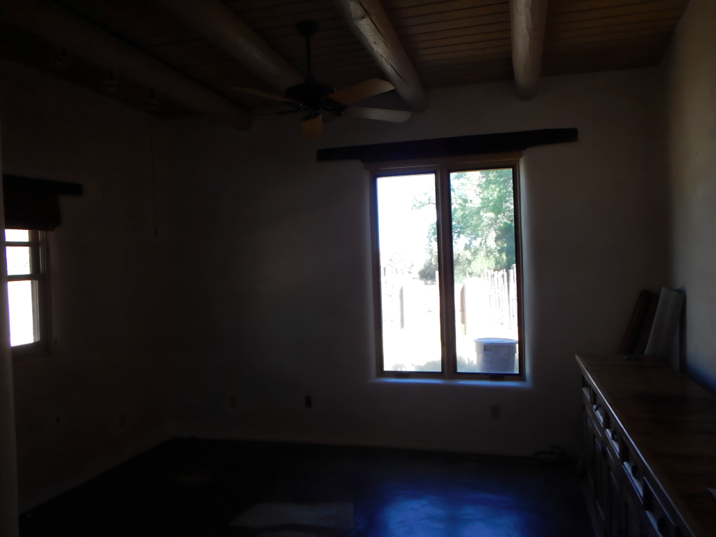 A dimly lit room with exposed wooden ceiling beams, a ceiling fan, and a large window that brings in natural light. The space appears empty, with a bare floor and minimal furnishings, suggesting it's ready for renovation or decoration.