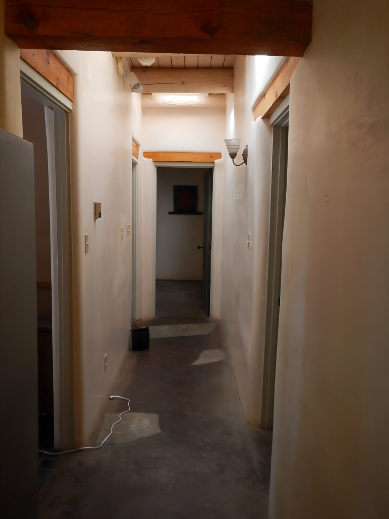 A narrow hallway with rustic plaster walls and wooden beams overhead, leading from the kitchen to the master bedroom. The simple, unfinished flooring and minimal lighting create a cozy yet slightly dated atmosphere in this passageway.