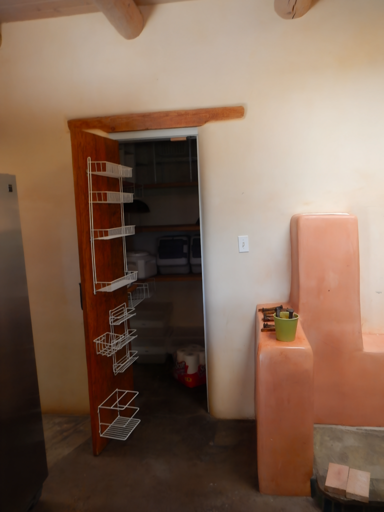 This image features a kitchen with a unique corner structure, a door with a hanging rack, and an adjoining pantry area. The smooth plastered walls and wooden beams create a warm atmosphere.