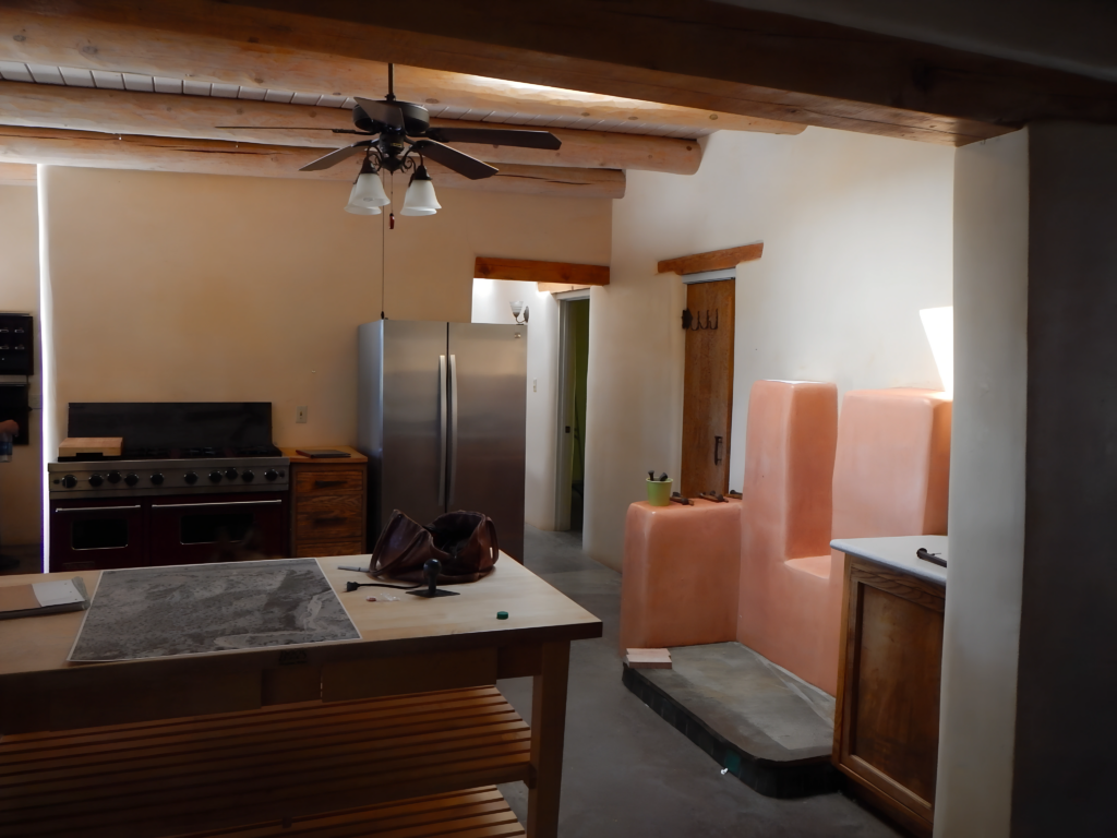 A rustic kitchen with wooden beams, a ceiling fan, and a large central island. The space features a stainless steel refrigerator, a traditional-style stove, and earthy-toned plaster elements, creating a warm, Southwestern-inspired atmosphere.