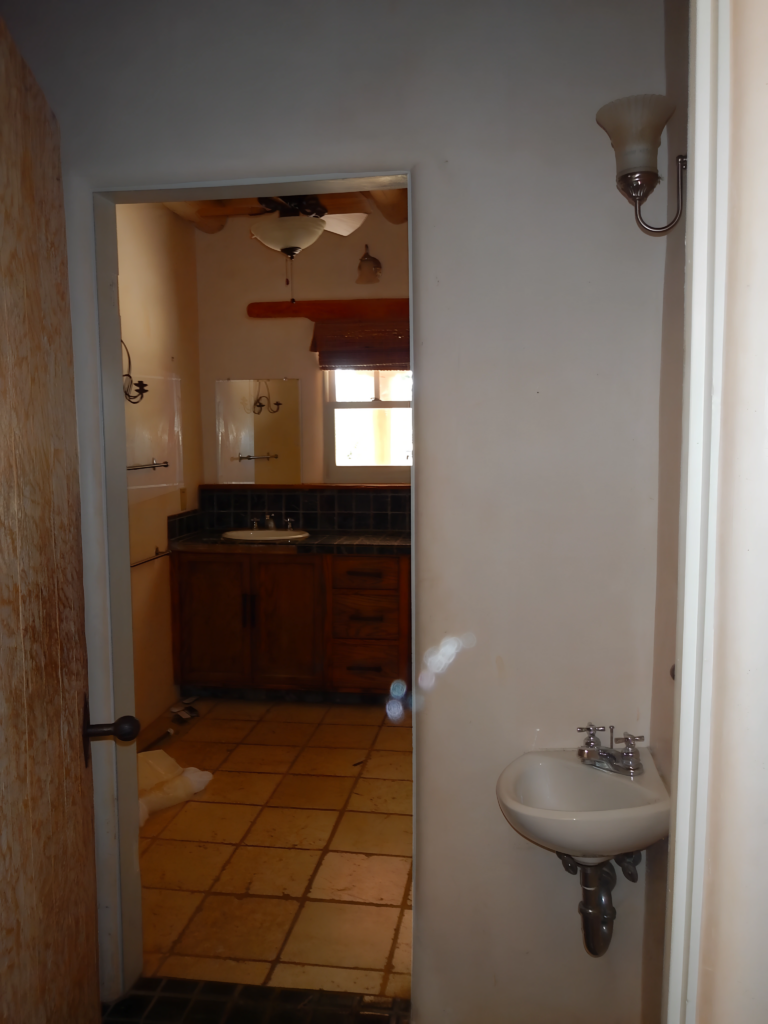 A photo of a bathroom before renovation, showing a small sink and a glimpse of the other area with a wooden cabinet and window in the background.