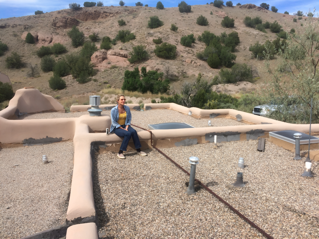 A person sitting on a rooftop, overlooking a scenic landscape with mountains, with architectural elements like rooftop vents and skylights visible.