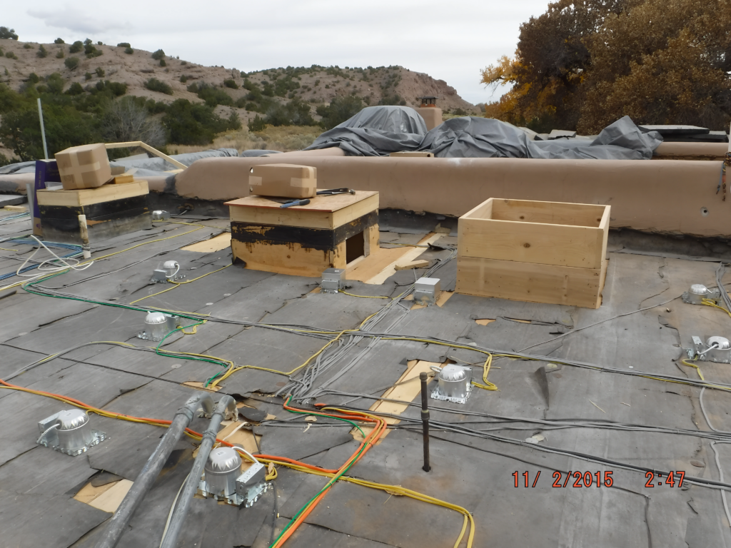 A rooftop under construction or renovation, with wooden structures, cables, and various equipment scattered across the surface. The surrounding desert landscape and trees are visible in the background, indicating an ongoing outdoor project in a remote area.