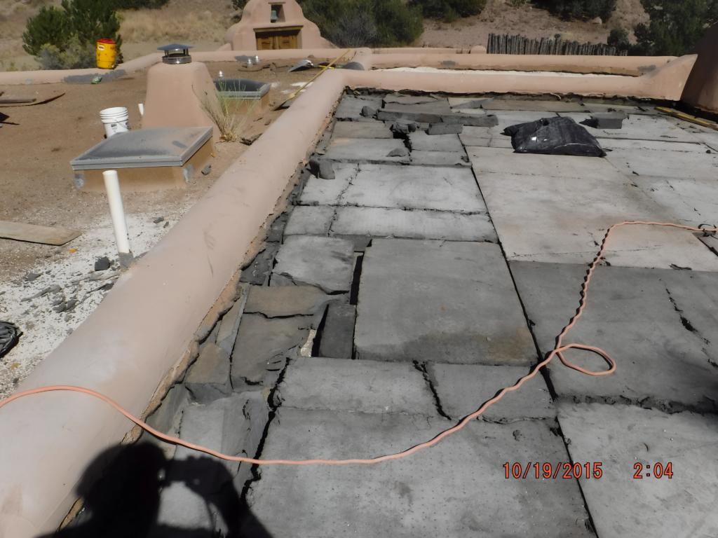 A rooftop undergoing repair or maintenance, with partially removed or damaged tiles and scattered tools. An orange extension cord lies across the surface, indicating ongoing work. The surrounding desert landscape and light-colored walls are visible in the background.