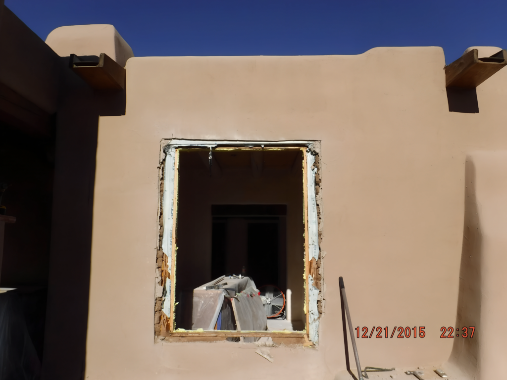 A partially renovated exterior wall with an open window frame, showing ongoing construction work. The surrounding plaster wall is intact, while tools and materials are visible inside the room, indicating preparations for further updates or installations.