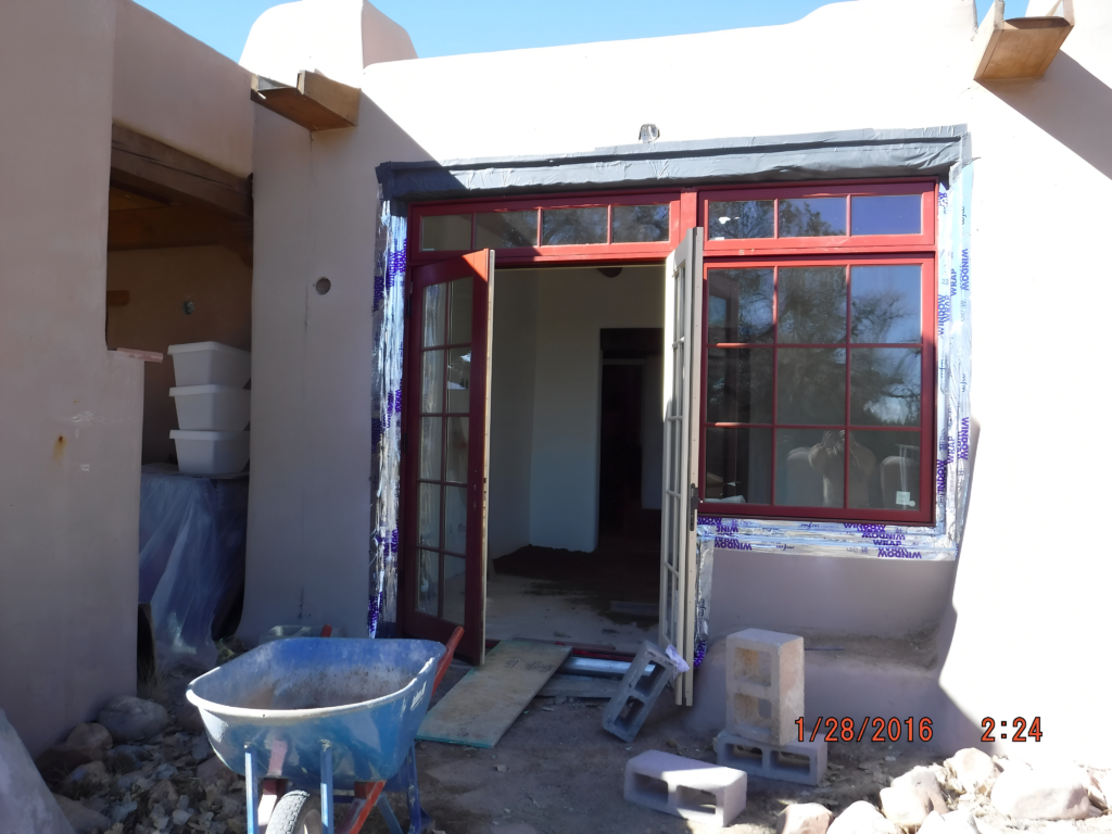 An exterior view of a room under renovation, featuring newly installed red-framed glass doors. Construction materials, including bricks and a wheelbarrow, are scattered outside, indicating ongoing work on the building and its surrounding area.