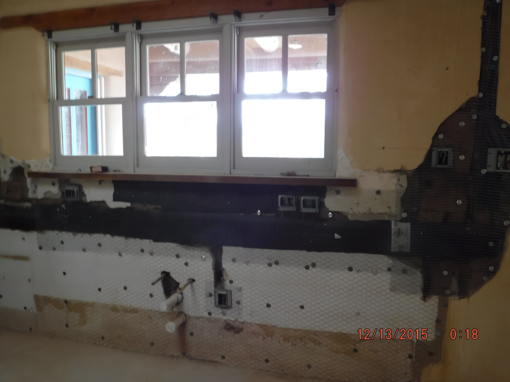 A kitchen under renovation with exposed walls and electrical wiring beneath a set of windows. The area is prepped for new installations, with missing cabinets and unfinished surfaces indicating ongoing remodeling work. Natural light enters through the windows.